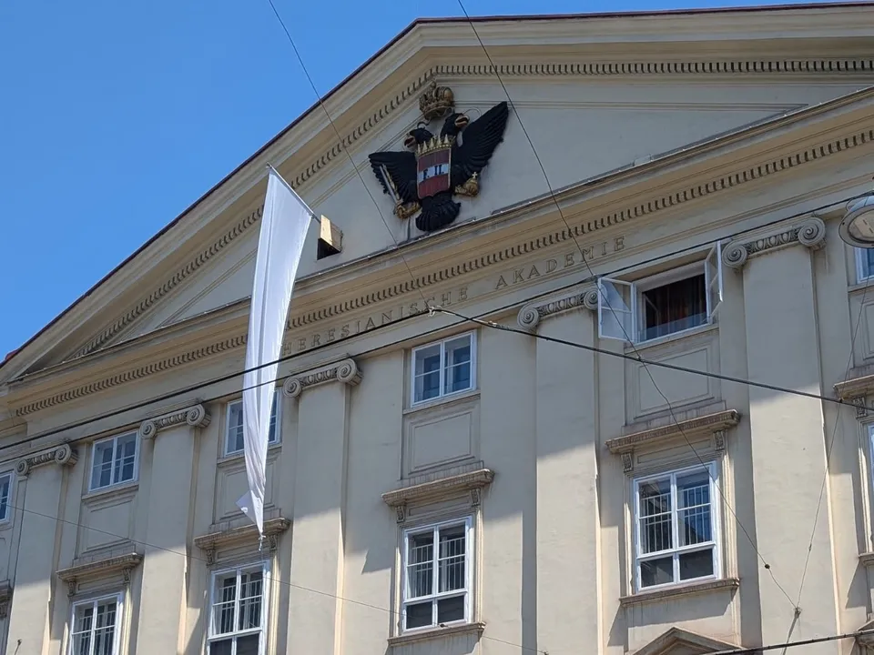 Aussenansicht des Theresianums, einem historischen Gebäude mit Doppeladler und Giebeldach, an dem eine weiße Fahne weht.