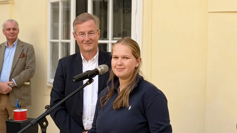 Ein Mann mit Brille, weißem Hemd und dunklem Sakko und eine junge Frau mit blonden langen Haaren vor einem Mikrofon stehend und in die Kamera blickend.