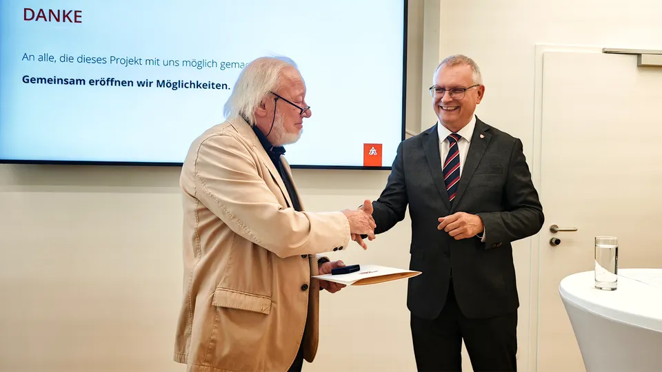 Shakehand von zwei lachenden Männern im Anzug vor einem großem Bildschirm stehend.