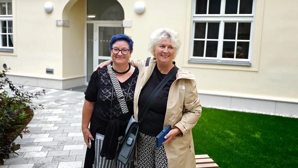 Zwei Frauen Arm in Arm in einem Gebäudeinnenhof stehend und in die Kamera lachend.