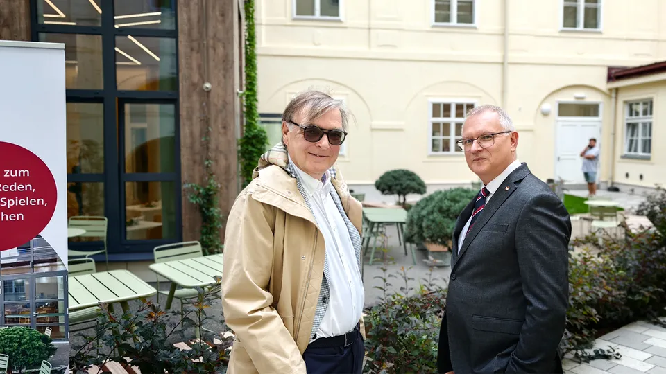 Zwei Männer in Anzug in einem Gebäudeinnenhof in die Kamera lachend.