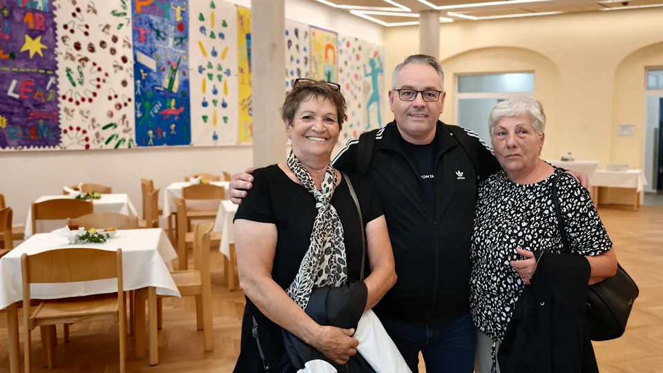Ein Mann mit zwei Frauen im Arm in einem Speisesaal mit bunten Hintegrundbildern stehend.