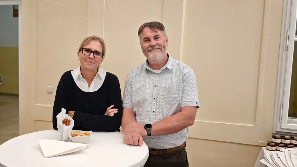 Ein Mann und eine Frau an einem Bistrotisch stehend und in die Kamera lachend.