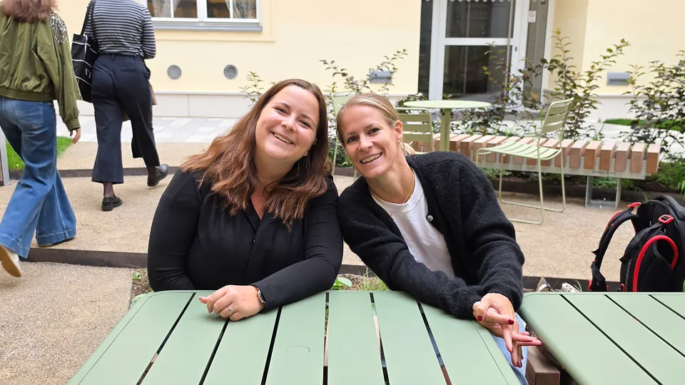 Zwei junge Frauen im Freien an einem grünen Tisch sitzend und in die Kamera lachend.