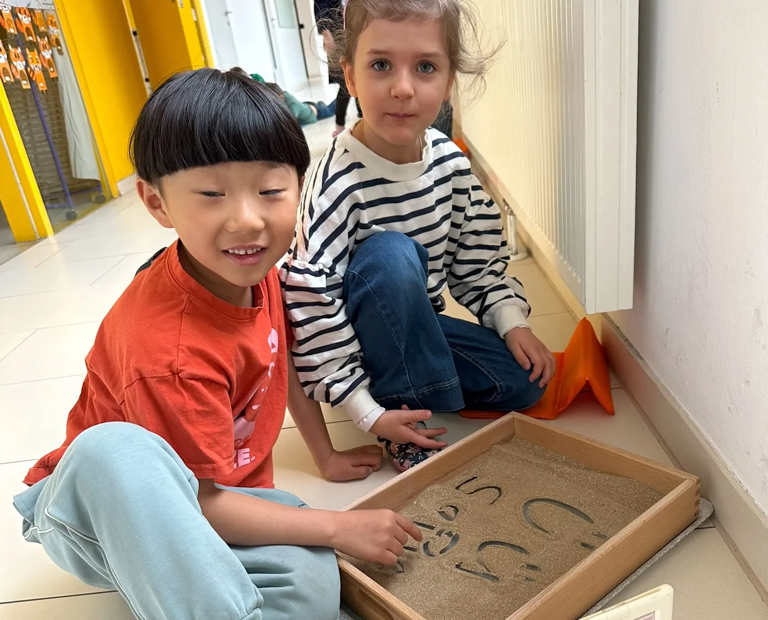Zwei Kinder am Boden sitzend mit einer kleinen Sandkiste vor sich, in der sie Buchstaben gezeichnet haben. Der Bub und das Mädchen blicken beide freundlich in die Kamera.
