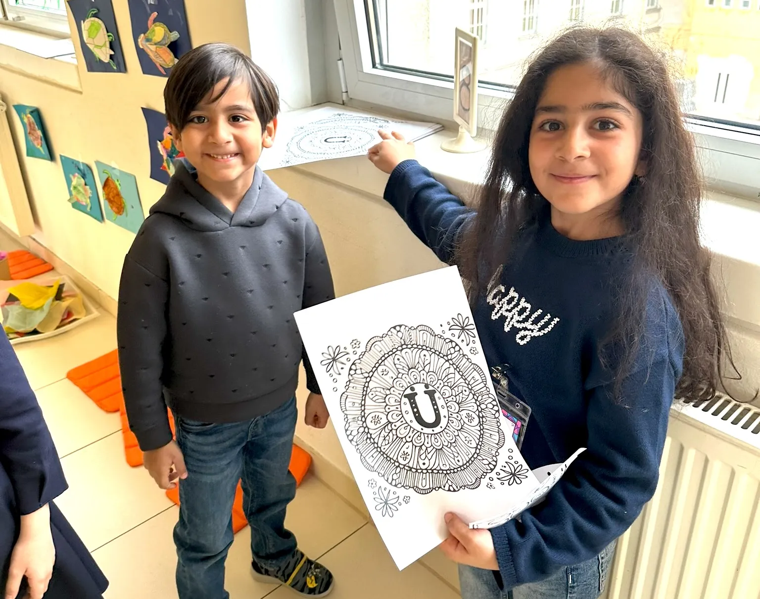 Zwei Schulkinder in die Kamera lachend, das Mädchen mit langen Haaren hält ein Mandala in die Kamera, der etwas jüngere Bub lacht fröhlich.