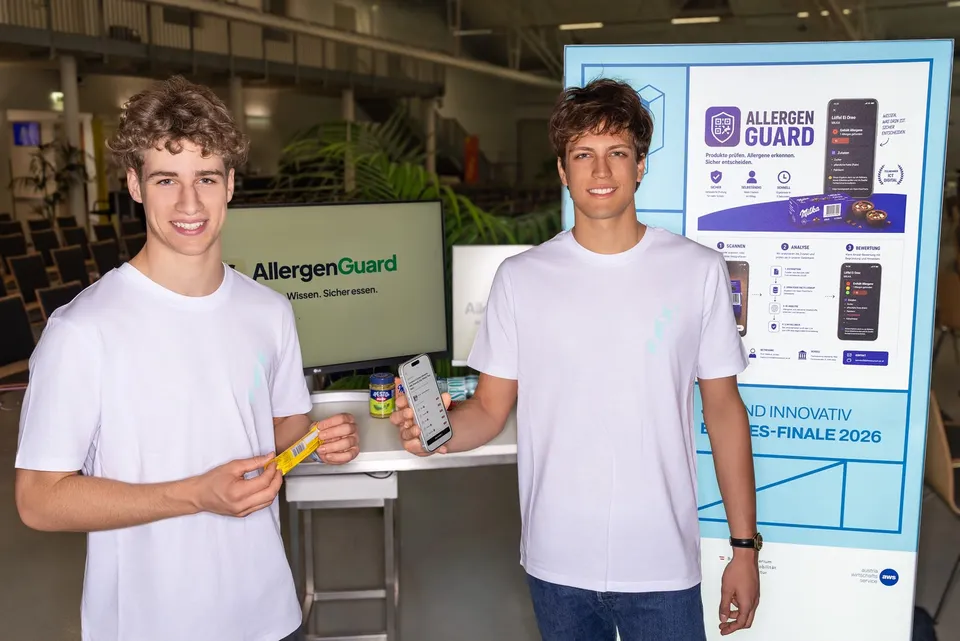 Zwei junge Burschen mit weißen T-Shirts auf einem Messestand vor einem Bildschirm stehend und eine Handy App präsentierend