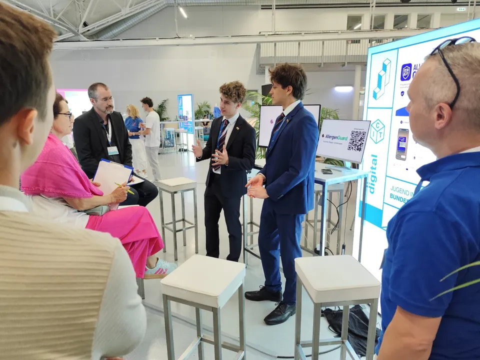 Messestand mit zwei jungen Burschen in Anzügen, die Besuchern eine App erklären.