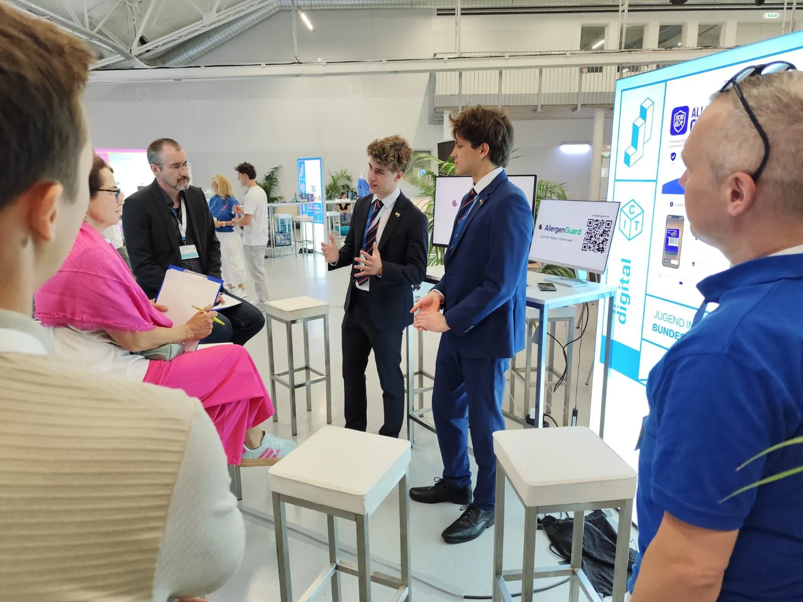 Messestand mit zwei jungen Burschen in Anzügen, die Besuchern eine App erklären.