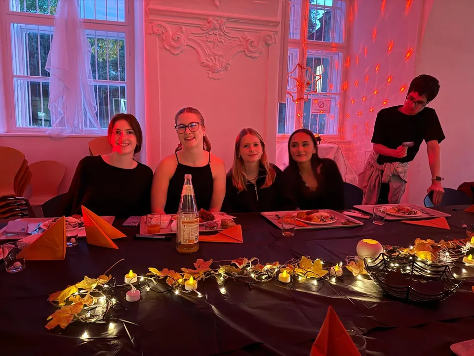 Five people sitting at a dimly lit table decorated with orange napkins, autumn leaves, and string lights, enjoying a meal.