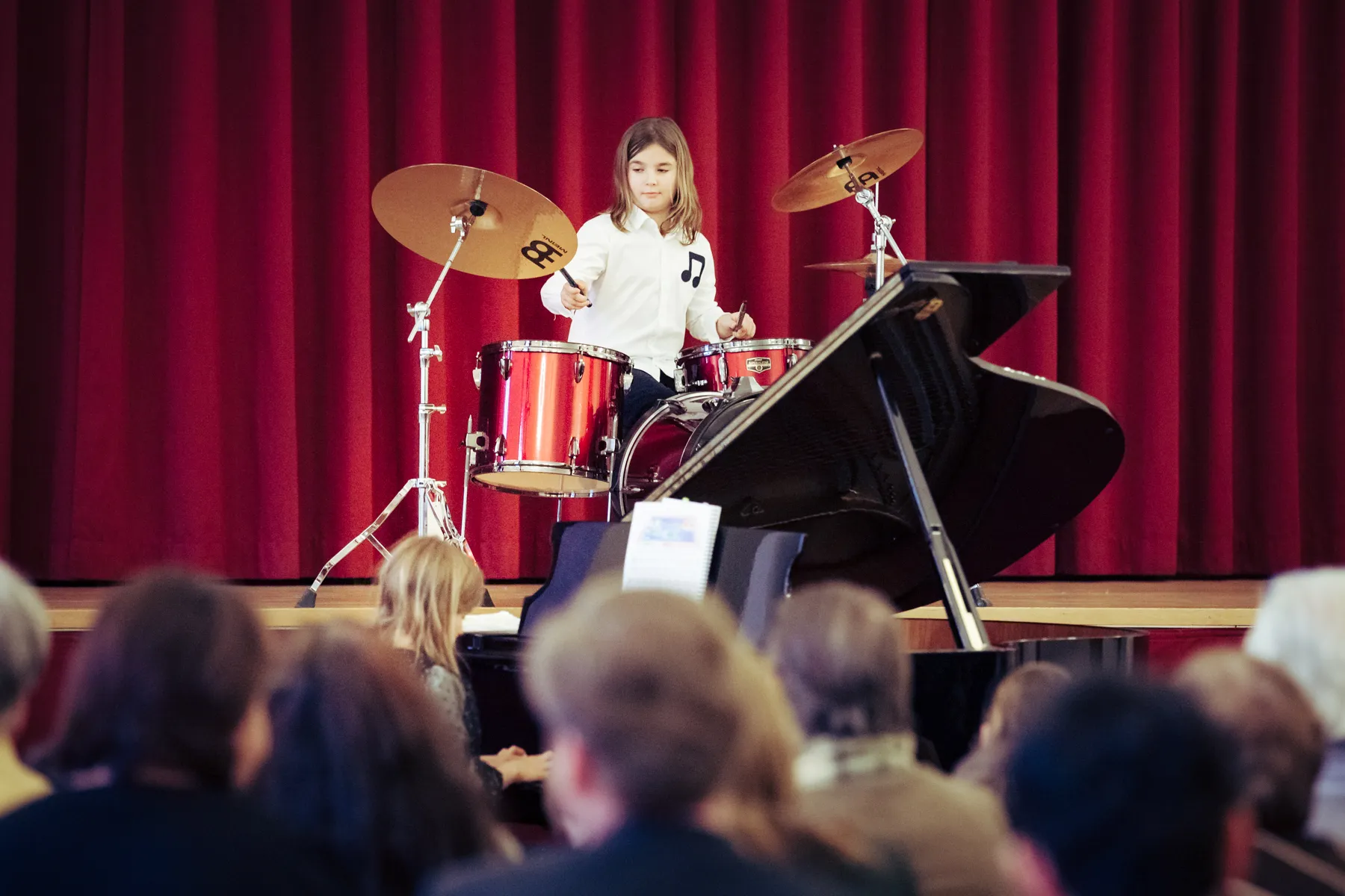 Mädchen auf einer Bühne vor rotem Vorhang Schlagzeug spielend mit Publikum im Vordergrund