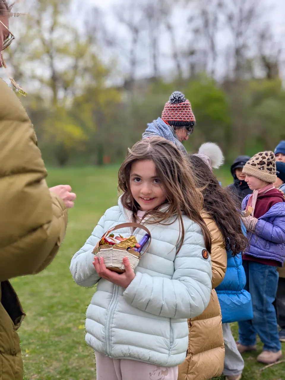 Mädchen mit braunen langen Harren und Winterkleidung stozlz ein Osterkörbchen in Händen haltend. Im Hintergrund eine erwachsene Frauen und weitere Kinder