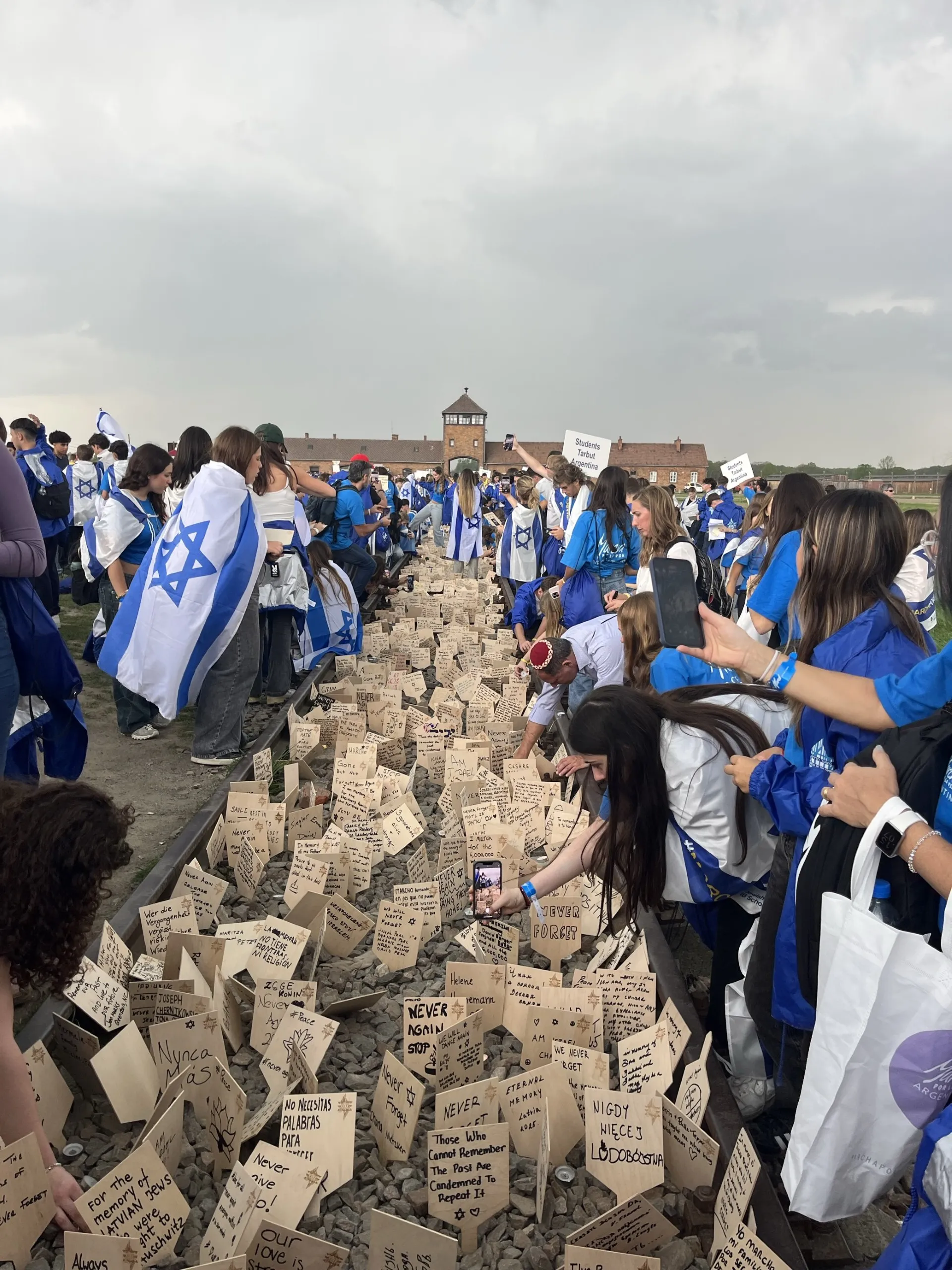 Gruppe junger Menschen mit jüdischen Flaggen um die Schultern gelegt entlang eines Beetes mit Steinen und Erinnerungskarten