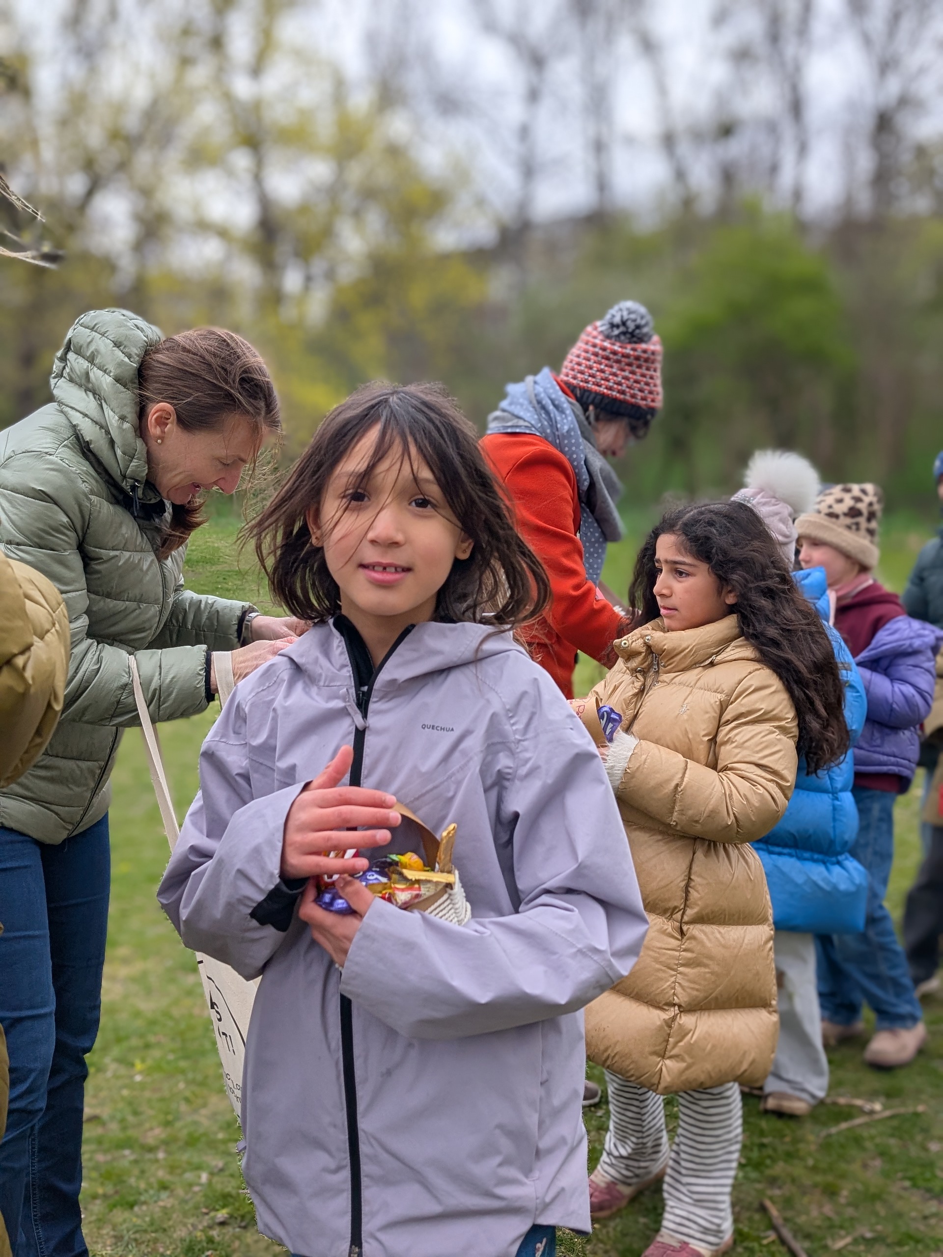 Mädchen mit dunklen langen Harren und ein Osterkörbchen in Händen haltend. Im Hintergrund zwei freundliche lachende erwachsene Frauen und weitere Kinder