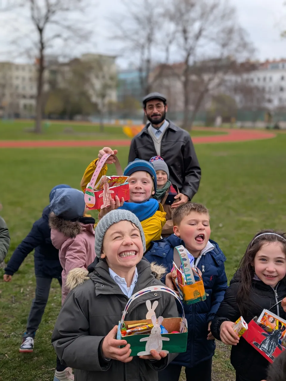 Gruppe von warm bekleideten Kindern fröhlich und grimassenschneidend ihre Osterkörbchen präsentierend. Im Hintergrund ein wohlwollend und zufrieden lachender erwachsener Mann mit Vollbart, Brille und Kappe