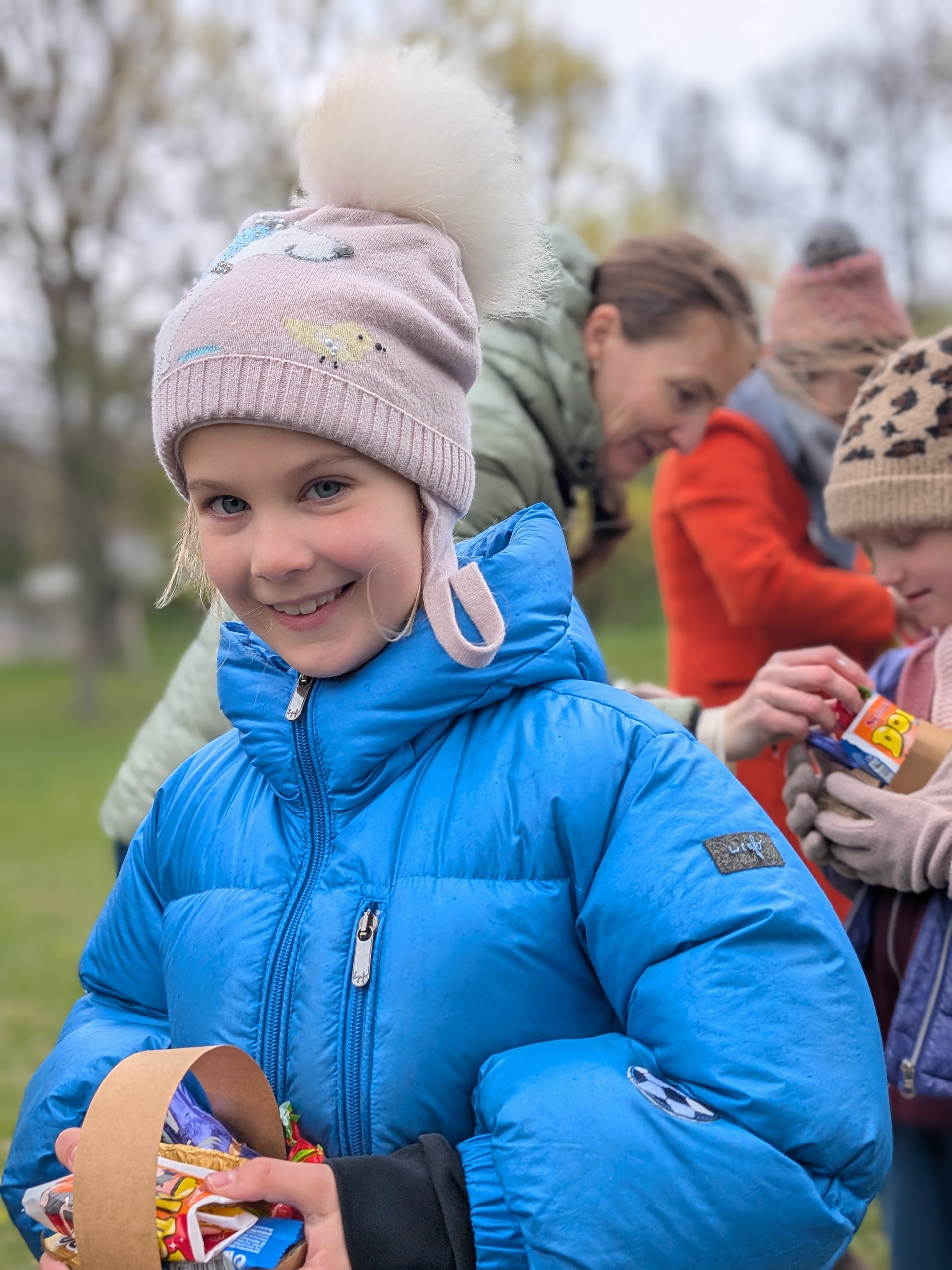 lachendes Mädchen mit Mütze und Winterkleidung ein Osterkörbchen stolze in Händen haltend. Im Hintergrund zwei freundliche lachende erwachsene Frauen und weitere Kinder