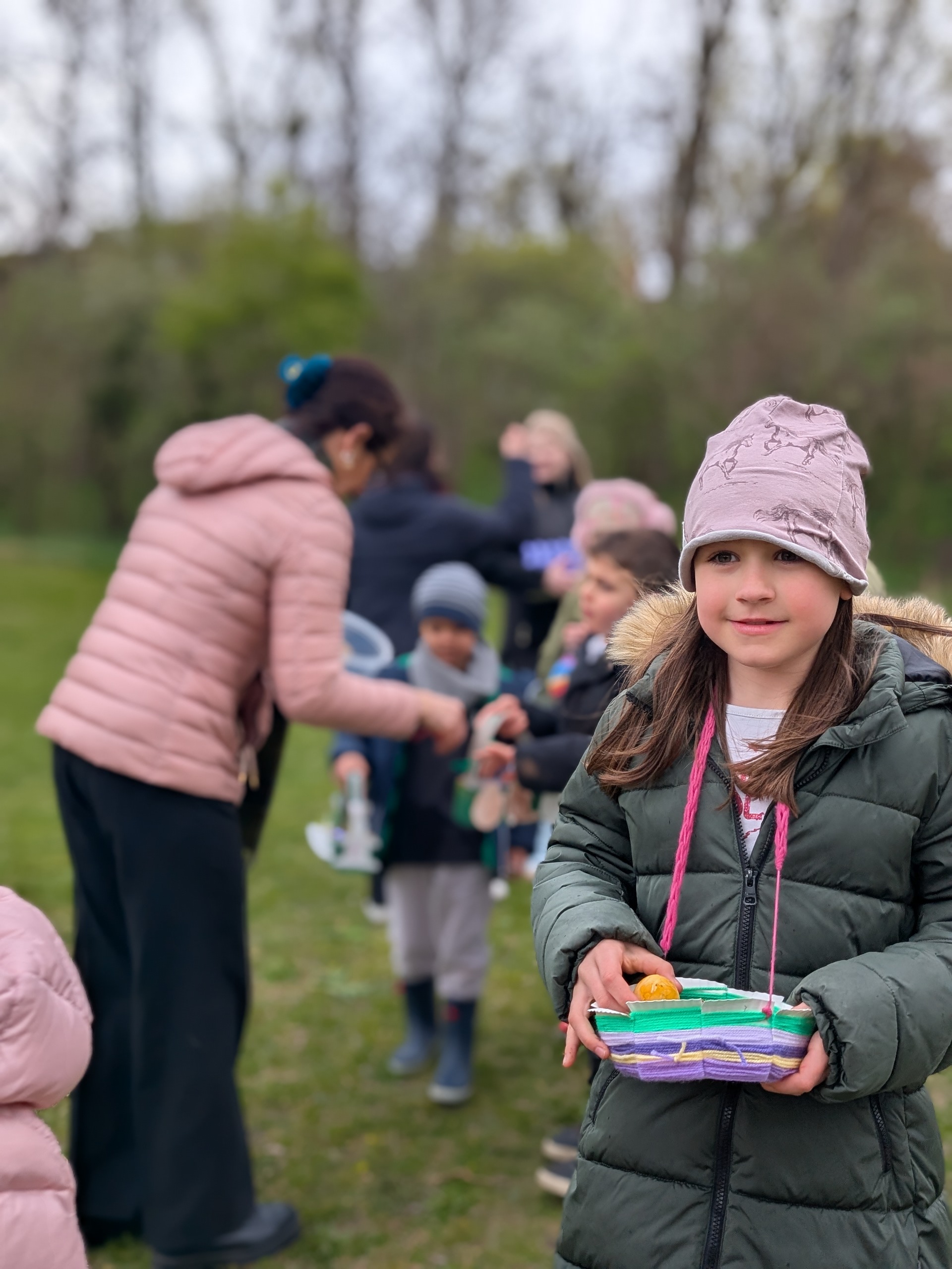 Mädchen mit lilafarbener Mütze, langen braunen Haaren und dunkelgrünem Parker und einem Osterkörbchen vor dem Körper mit einem orangenen Osterei in der Hand