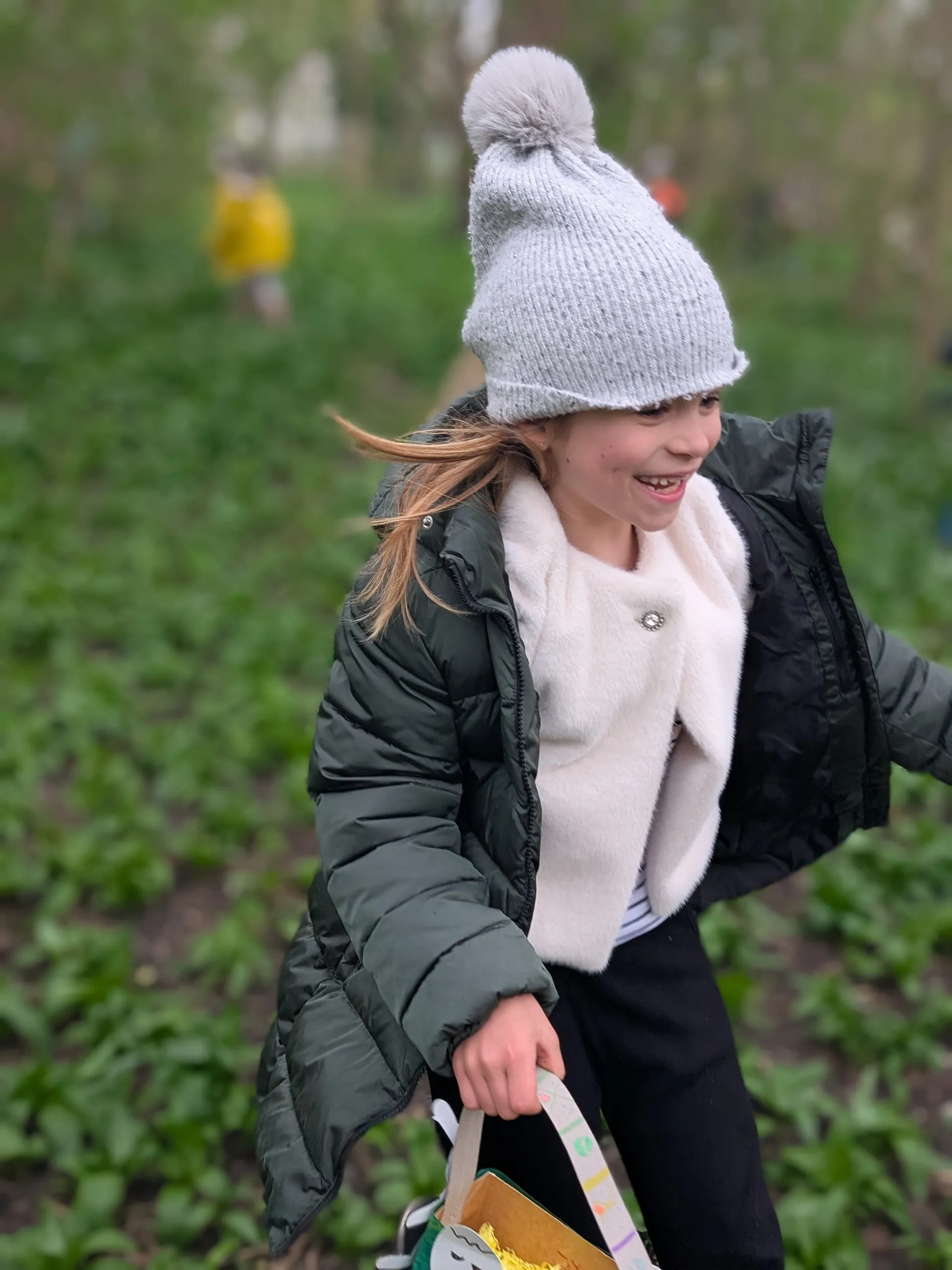 fröhlich lachendes Mädchen mit grau Haube, blonden langen Haaren und offenem Wintermantel läuft mit einem Osterkörbchen in der Hand durch ein Waldstück mit Bärlauch