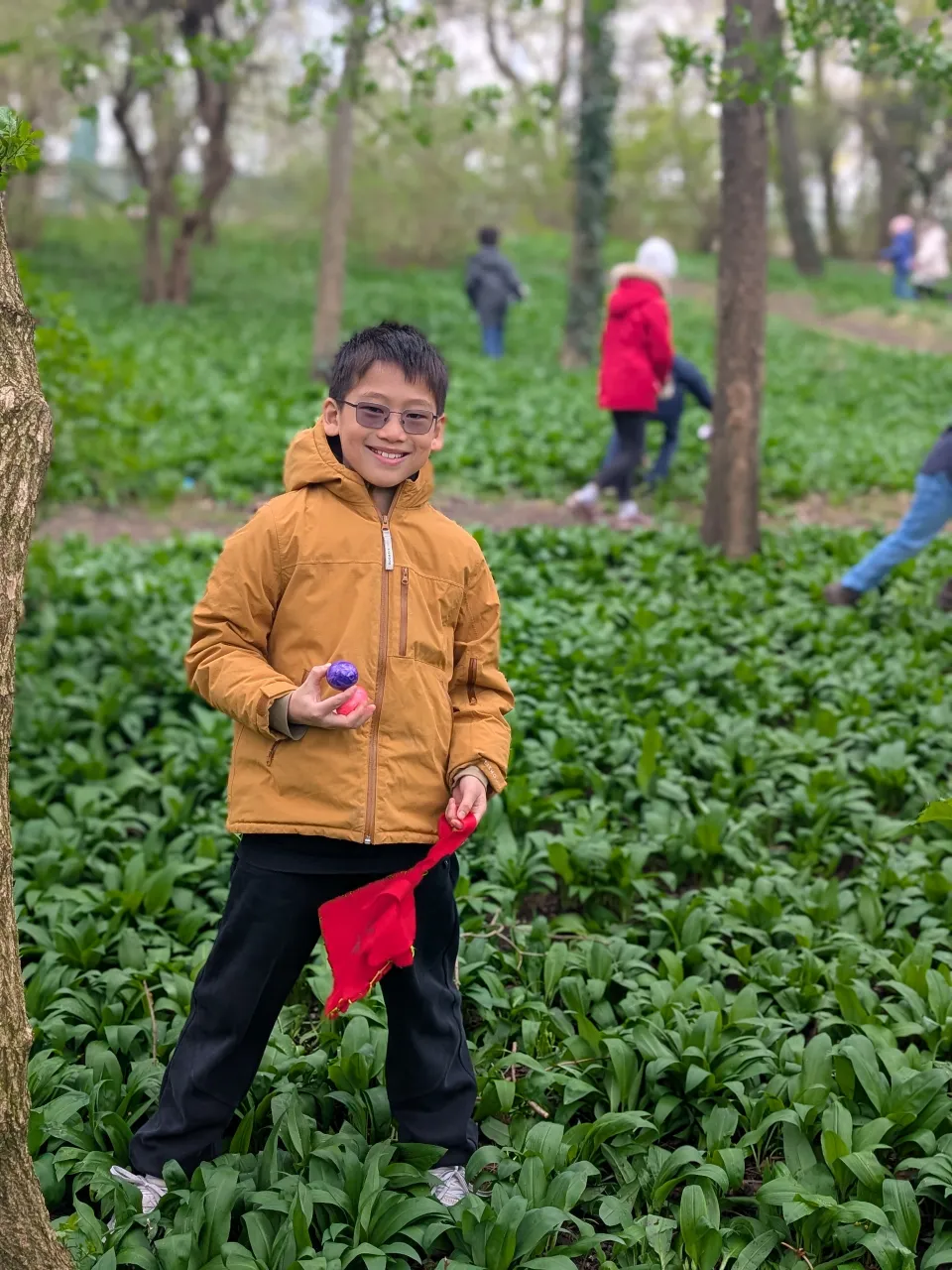 junger Bursche mit Brille und dunklen kurzen Haaren und einer ockerfarbenen Jacke hält freudestrahlend zwei Ostereier in der Hand. Hinter ihm laufen Kinder im bärlauchbedeckten Wald