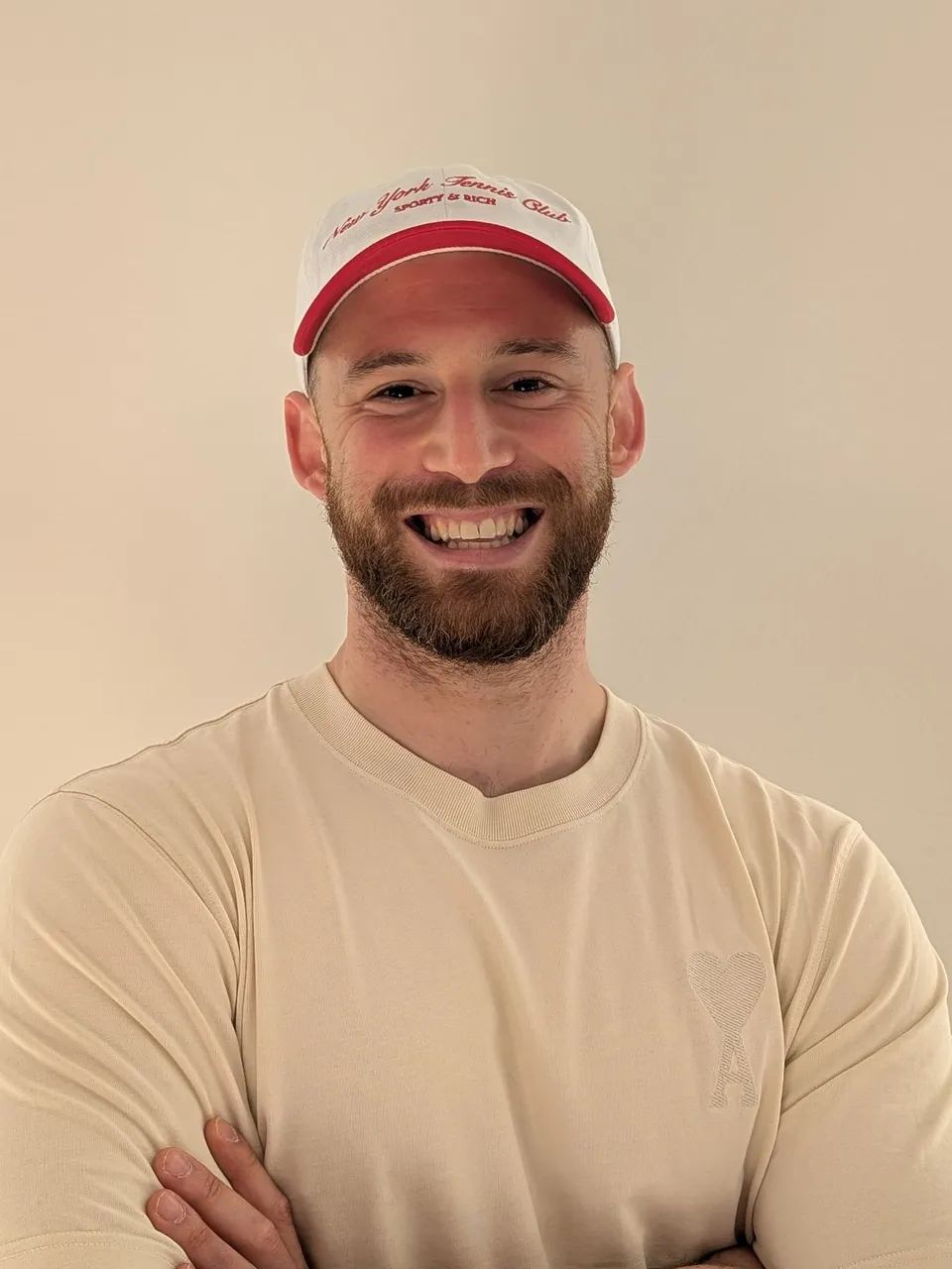 junger sportlicher Mann mit Baseballkappe und Vollbart in einem beigen T-Shirt und PowerPose in die Kamera lachend