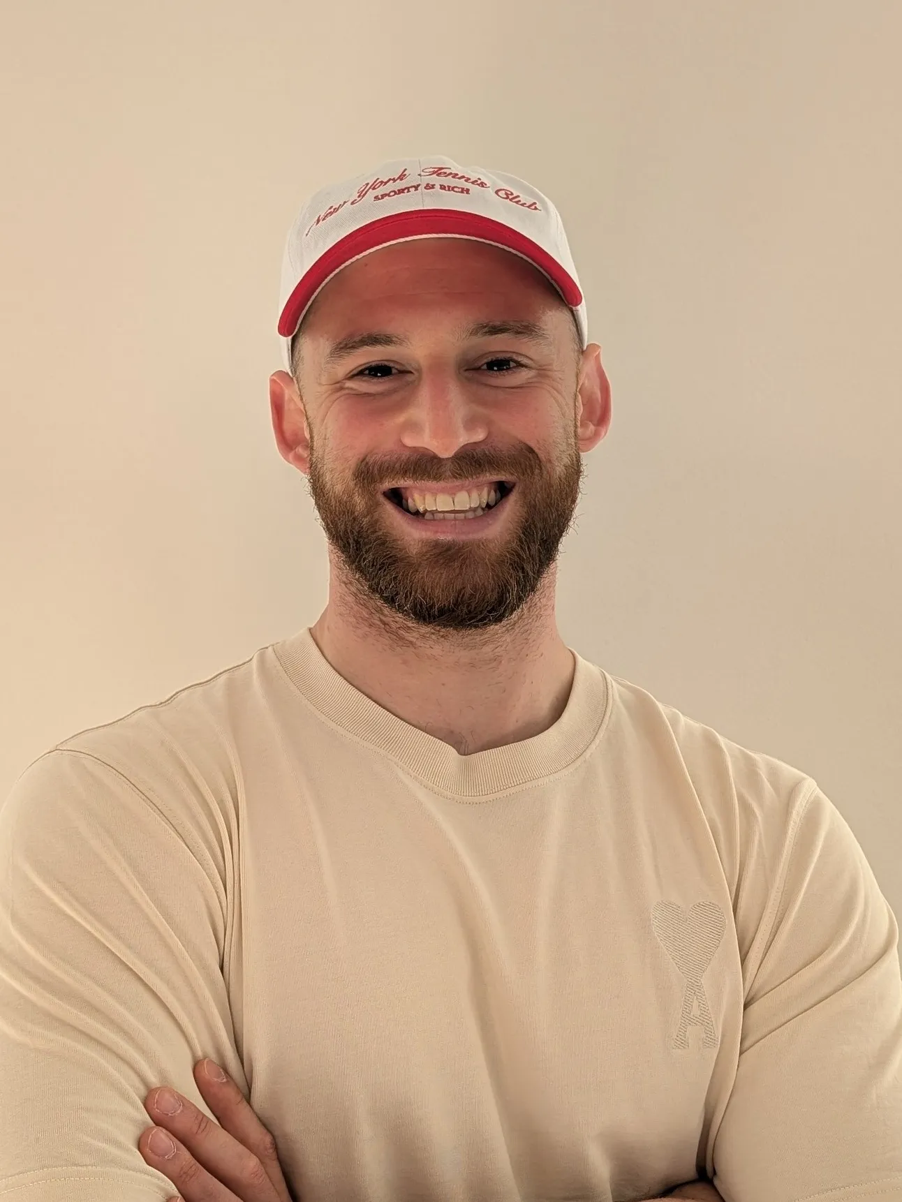 junger sportlicher Mann mit Baseballkappe und Vollbart in einem beigen T-Shirt und PowerPose in die Kamera lachend