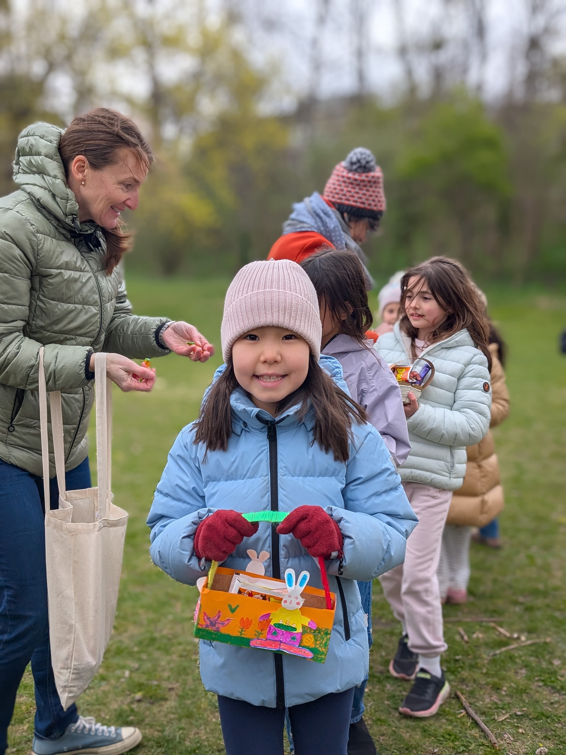 Mädchen mit dunklen langen Harren und Winterkleidung ein Osterkörbchen in Händen haltend. Im Hintergrund zwei freundliche lachende erwachsene Frauen und weitere Kinder