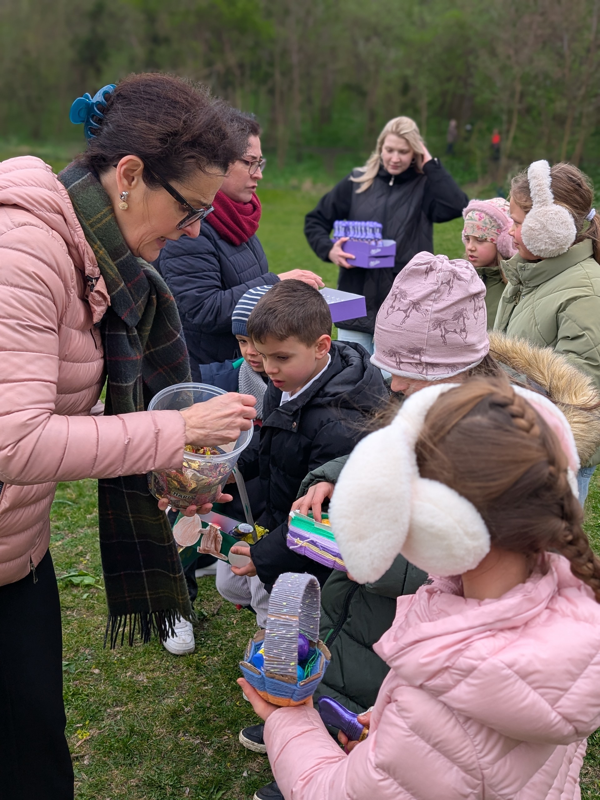 Gruppe von Kindern warm gekleidet vor einer erwachsenen eleganten Frau stehend, die den Kindern Süssigkeiten in ein Osterkörbchen gibt