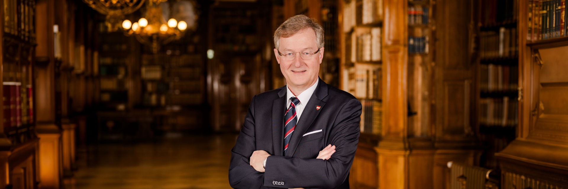 Mann in dunklem Anzug mit Krawatte, Stecktuch und Anstecknadel lächelnd in die Kamera blickend vor einem historischen Bibliothekshintergrund