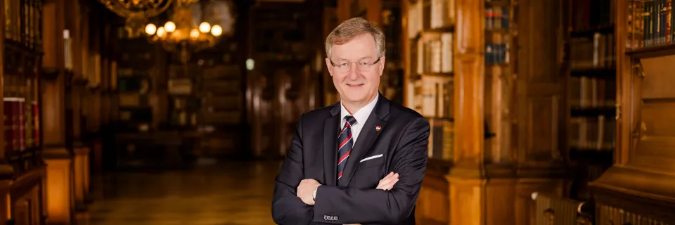 Mann in dunklem Anzug mit Krawatte, Stecktuch und Anstecknadel lächelnd in die Kamera blickend vor einem historischen Bibliothekshintergrund