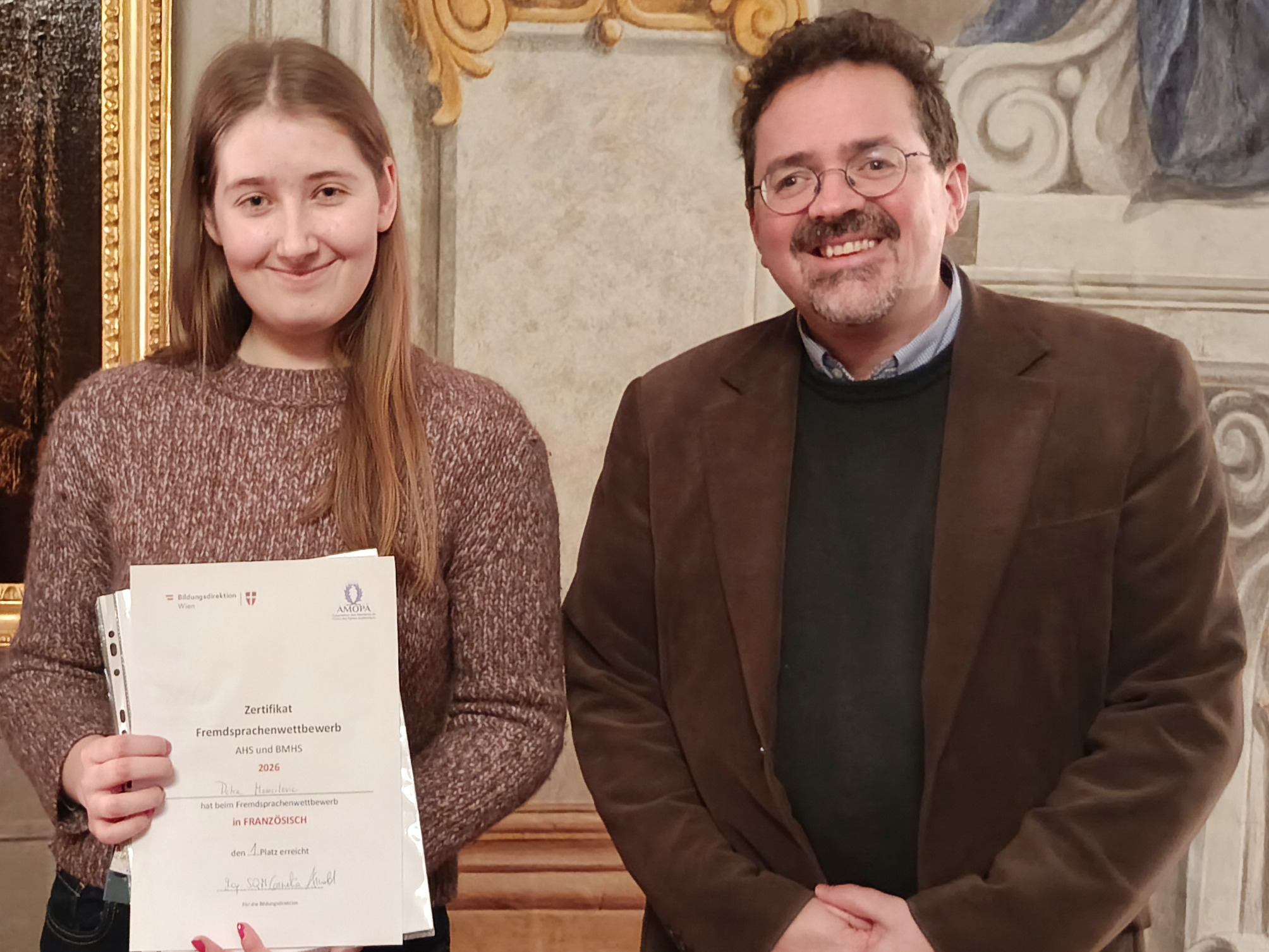 Eine junge Frau und ein 50jähriger Mann mit Brille und Bart in historischen Räumlichkeiten. Beide lachen in die Kamera. Die junge Frau hält ein Zertifikat in der Hand.