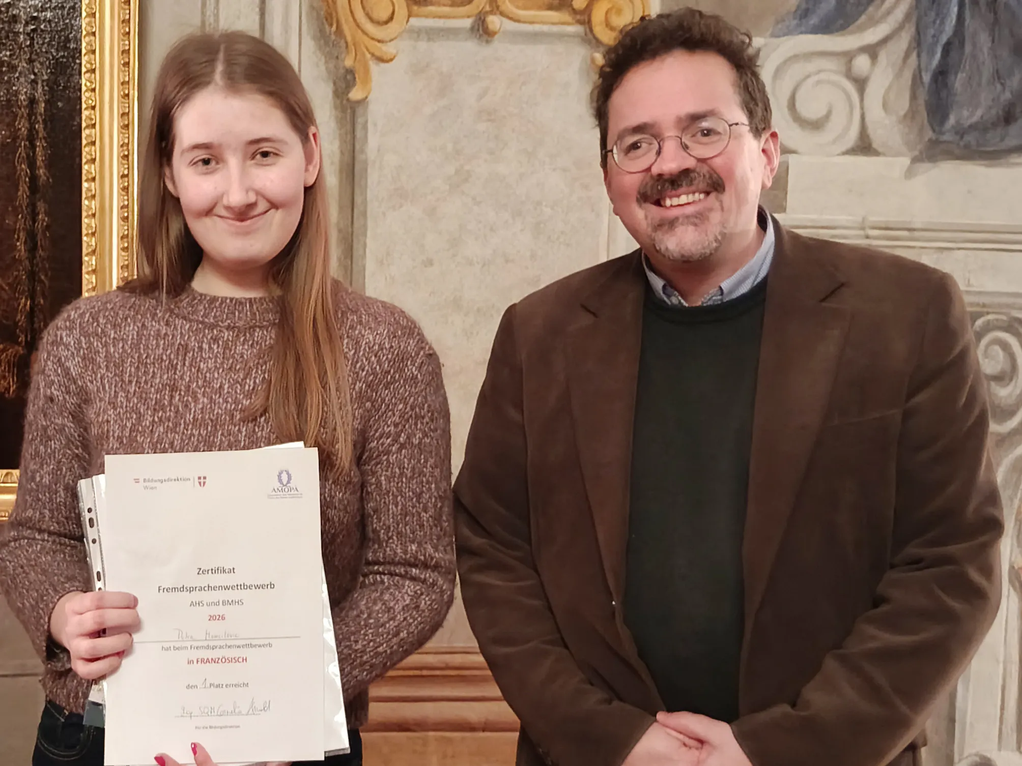 Eine junge Frau und ein 50jähriger Mann mit Brille und Bart in historischen Räumlichkeiten. Beide lachen in die Kamera. Die junge Frau hält ein Zertifikat in der Hand.