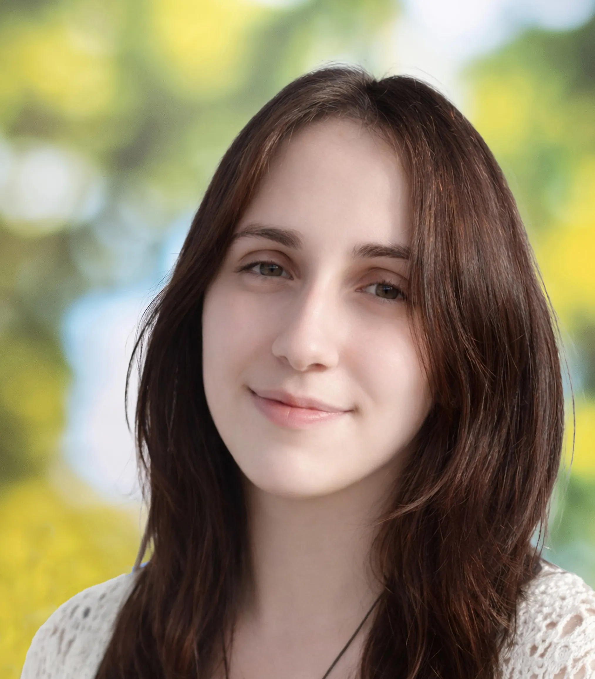 Woman with long brown hair smiling softly, wearing a white top, against a blurred background of green and yellow foliage.