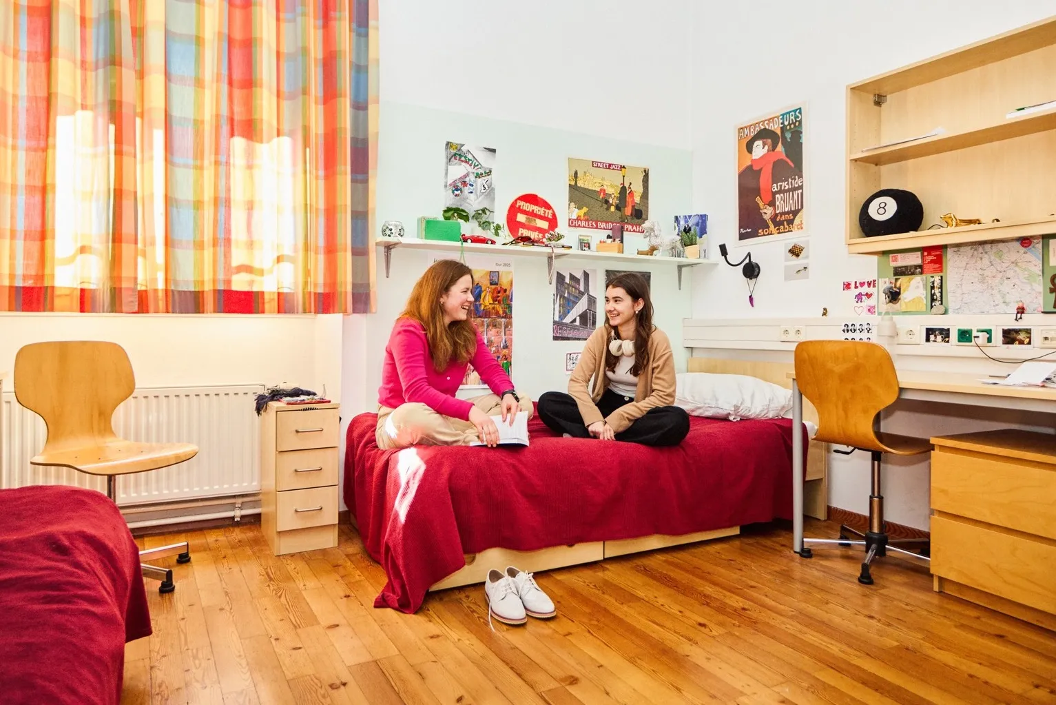 Zwei Mädchen in einem Jugendzimmer mit bunter Einrichtung auf einem Bett sitzend und miteinander plaudernd