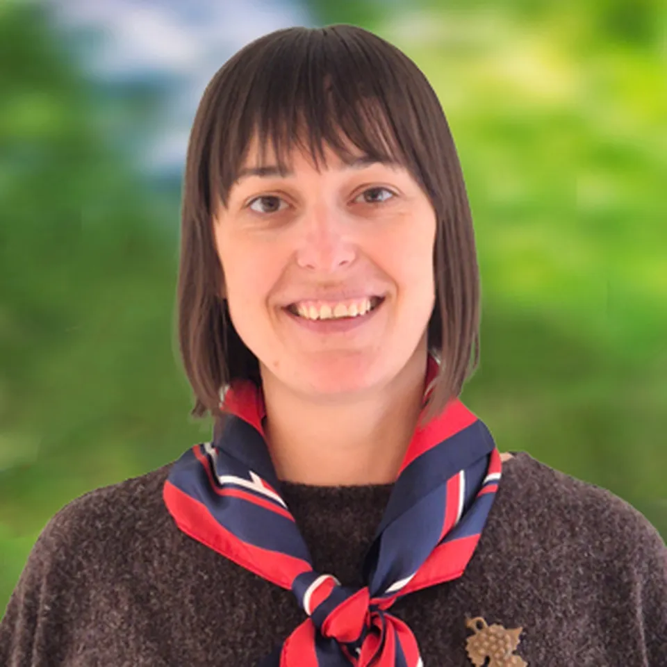 Lächelnde Frau mit schulterlangem Haar, die ein rot‑blaues Tuch und einen dunklen Pullover trägt.