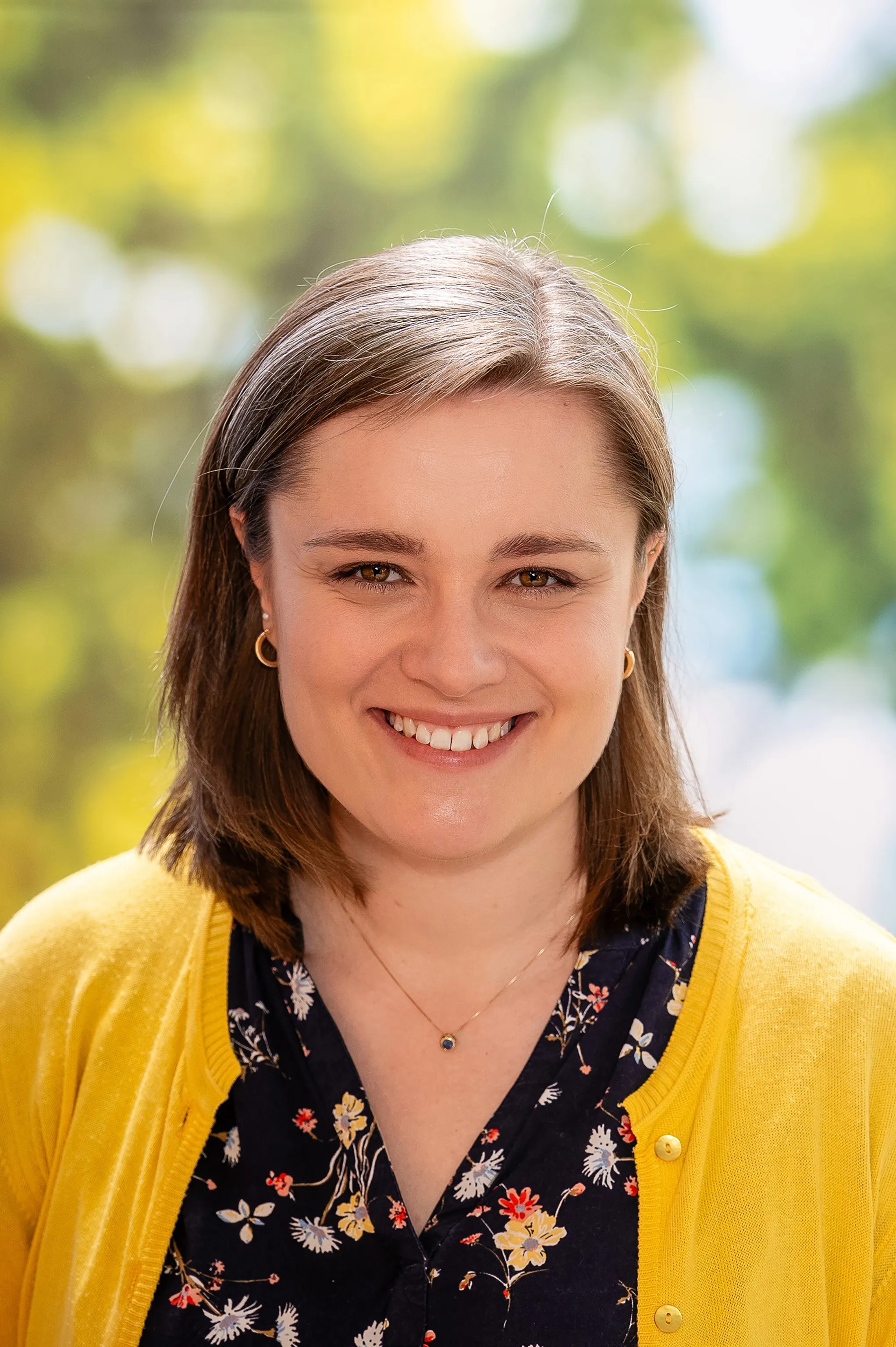 Lachende Frau mit kurzem braunen Haar, eine gelbe Weste und eine Blumenbluse tragend.