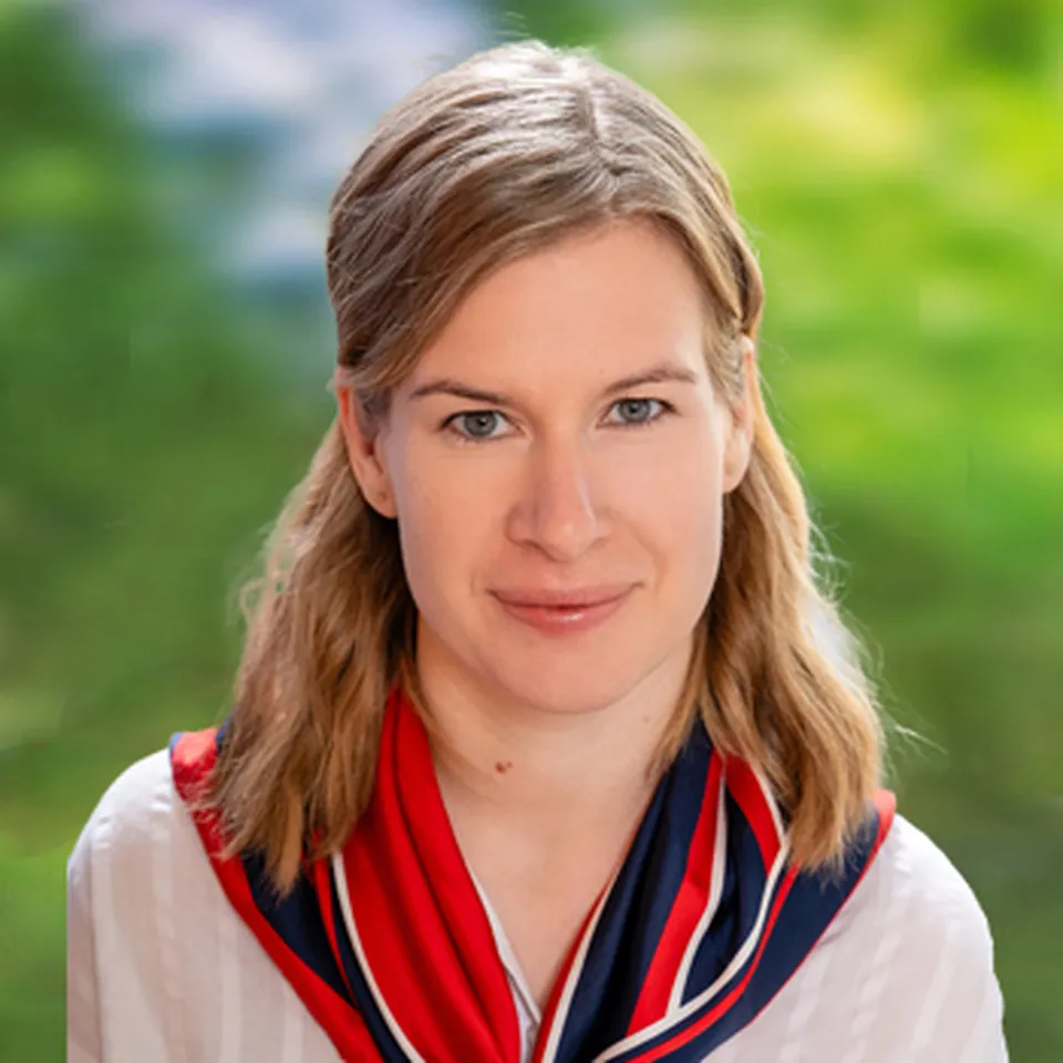 Woman with light brown hair, wearing a red and blue scarf over a white shirt, smiles softly with a blurred green and yellow background.