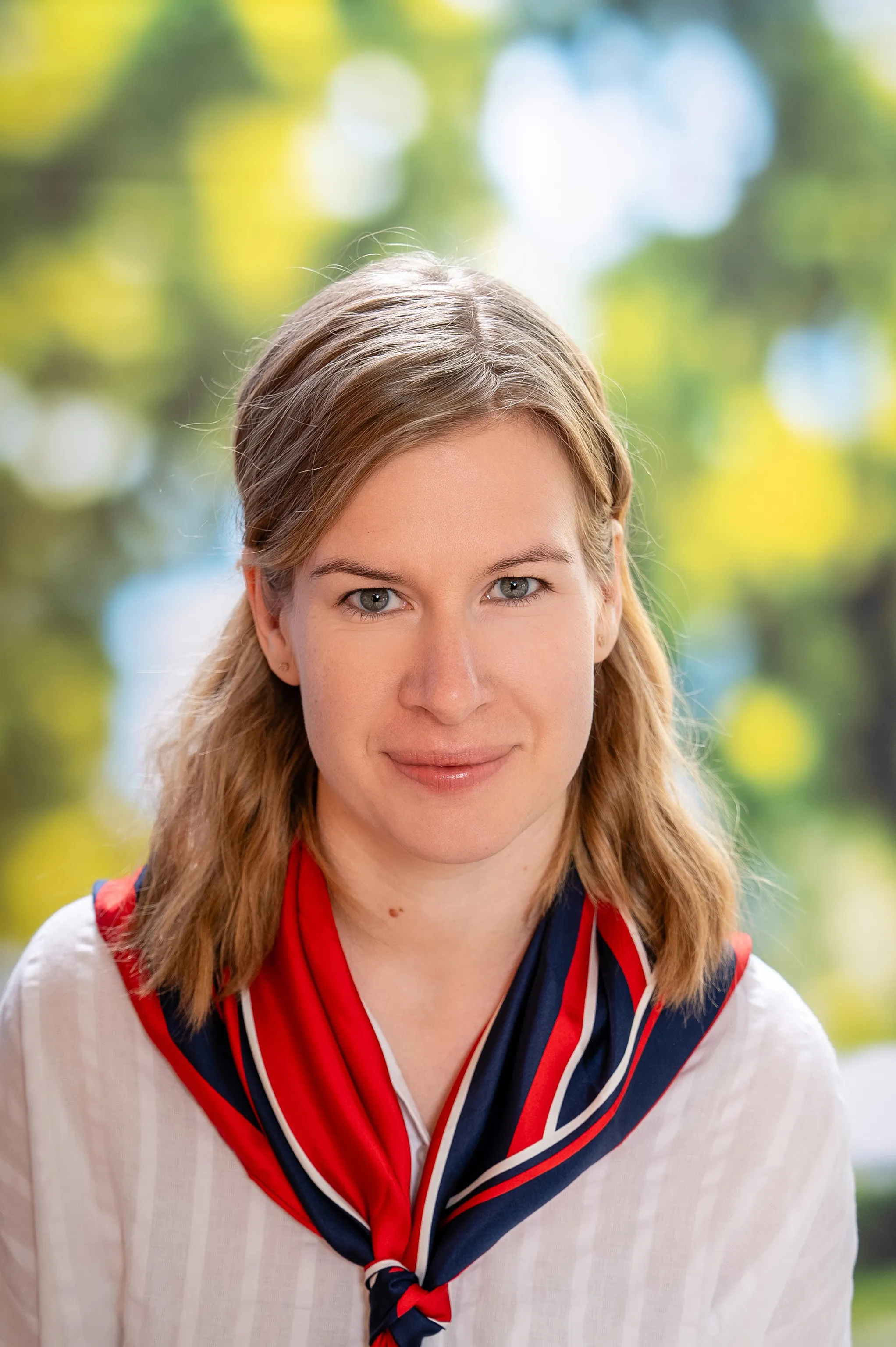 Woman with light brown hair, wearing a red and blue scarf over a white shirt, smiles softly with a blurred green and yellow background.