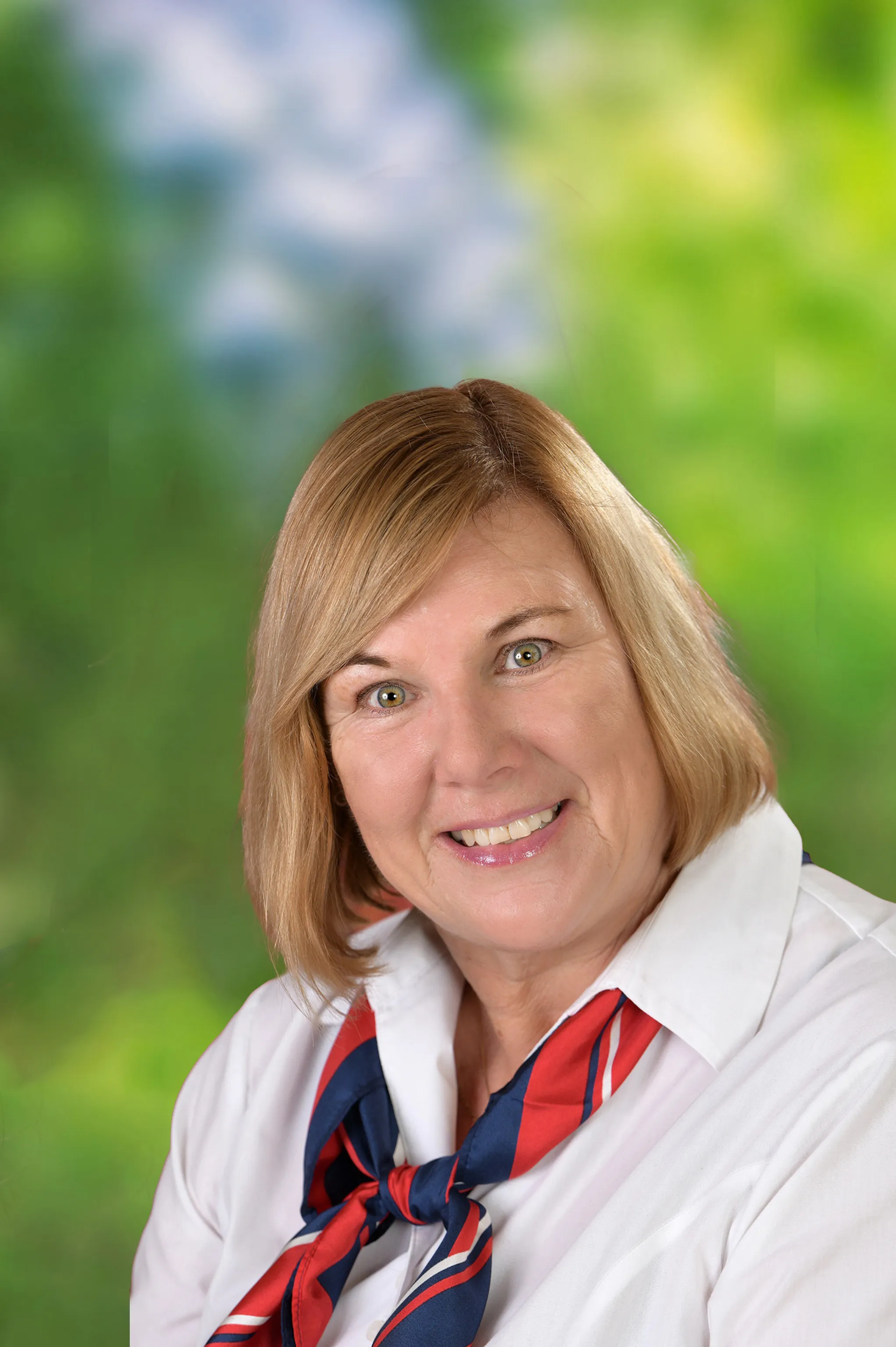 Lächelnde Frau mit blonden kurzen Haaren, die eine weiße Bluse mit einem rot-blauen Tuch trägt.