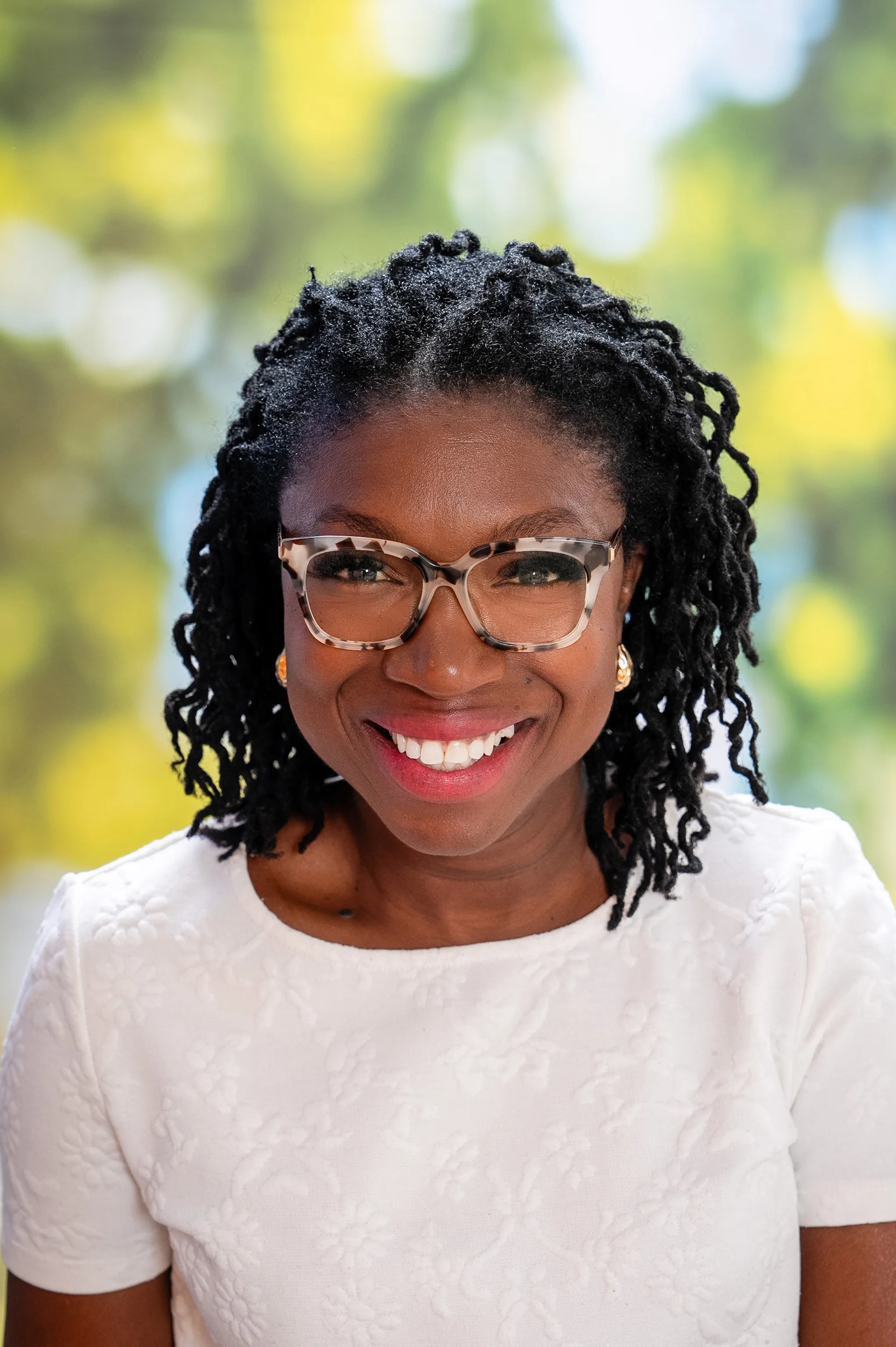 Lachende Frau mit lockigem Haaren und Brille, trägt ein weißes gemustertes T Shirt.