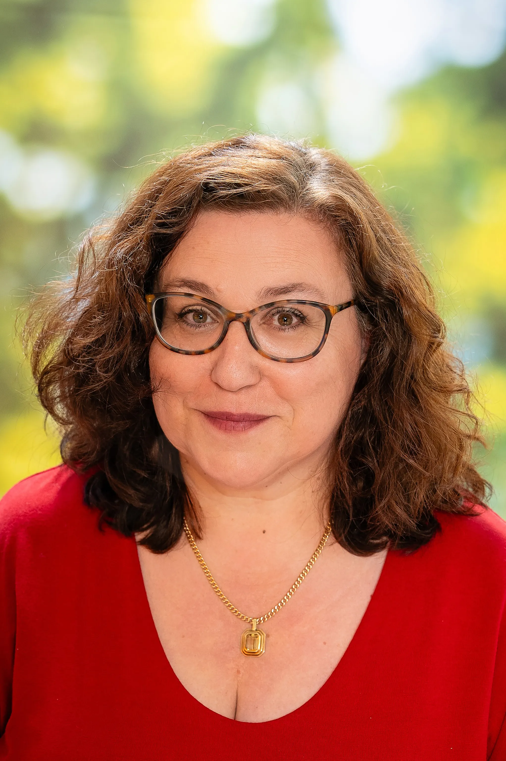 Eine Frau mit braunem gelpcktem Haar, Brille, einen roten Pullover und einer Halskette.