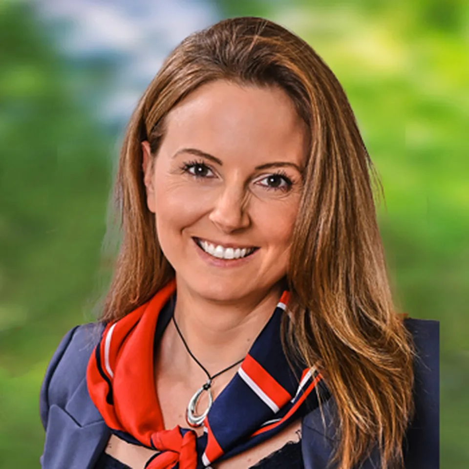 Lächelnde Frau mit langen braunen Haaren, mit eleganter dunkelblauer Jacke und rot‑blauesmTuch.