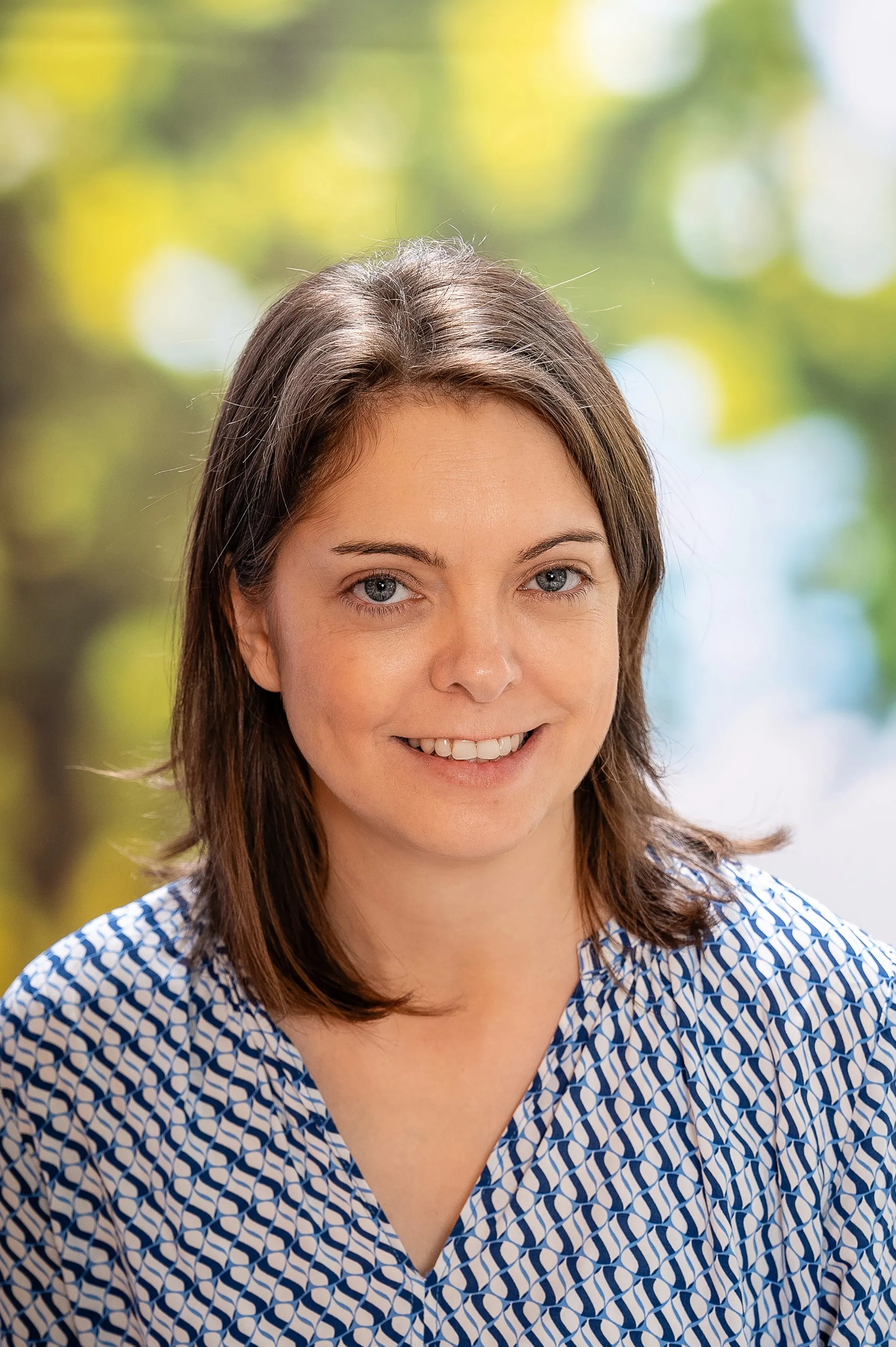 Eine lächelnde Frau mit braunen kurzen Haaren in einer blau gemusterten Bluse.