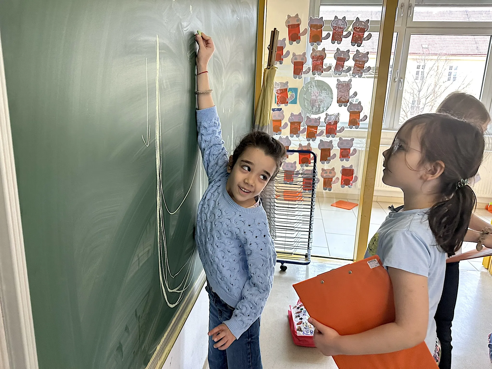 Zwei Mädchen bei einer Schultafel stehend, eines der Mädchen zeichnet mit einer Kreide einen Buchstaben an der Tafel nach, das andere Mädchen beobachtet sie dabei.