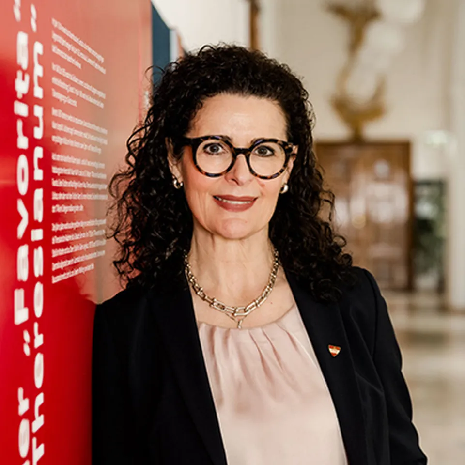 Frau mit dunkler Brille und glatt nach hinten frisierten dunklen Haaren mit eleganter dunkelblauer Jacke und rot blau weißem Seidentuch