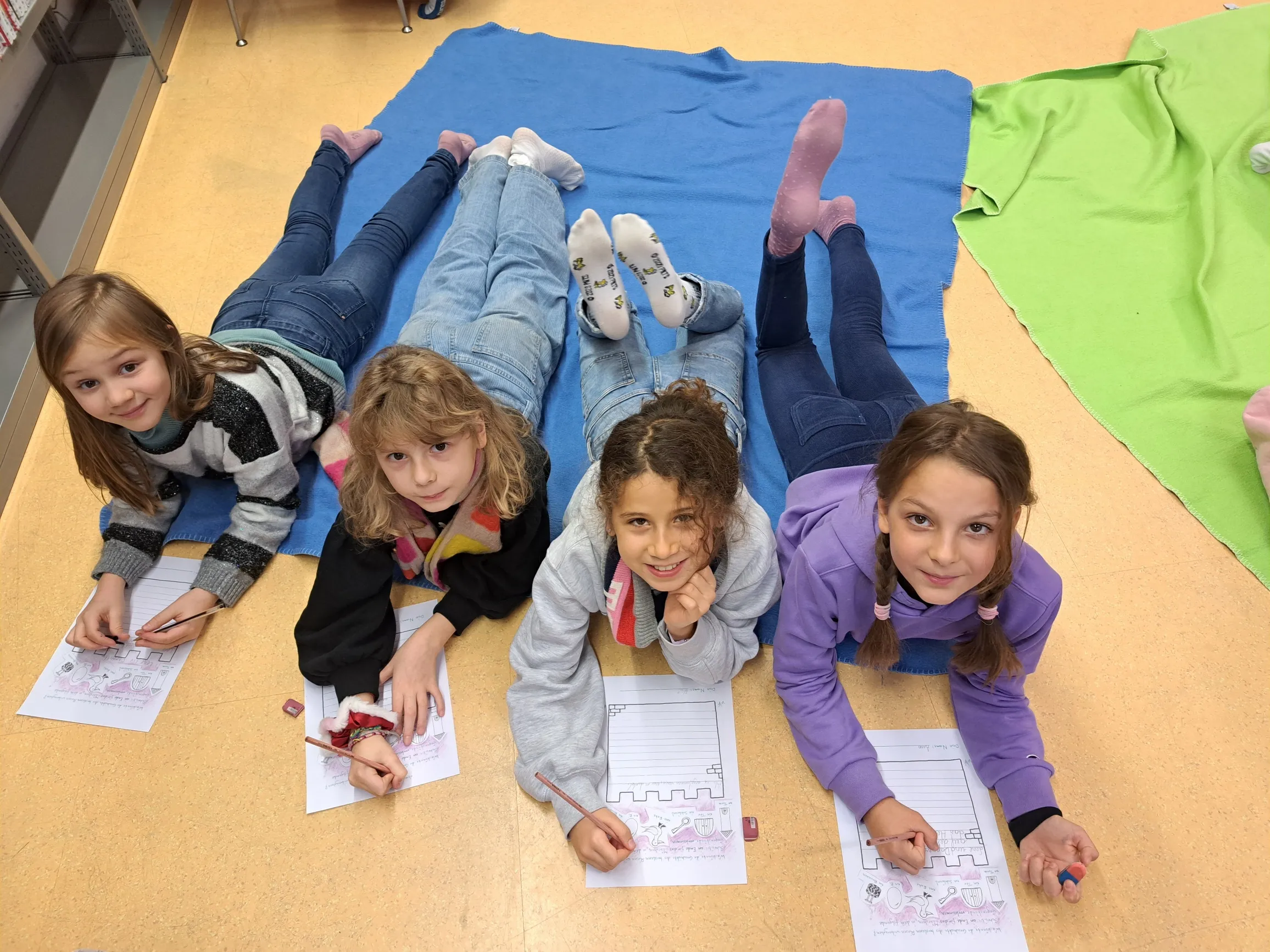 Vergnüngte Gruppe von vier Mädchen am Bauch am Boden liegend mit dem Kopf nach oben blickend, jede mit einem Arbeitsblatt und einem Stift in der Hand