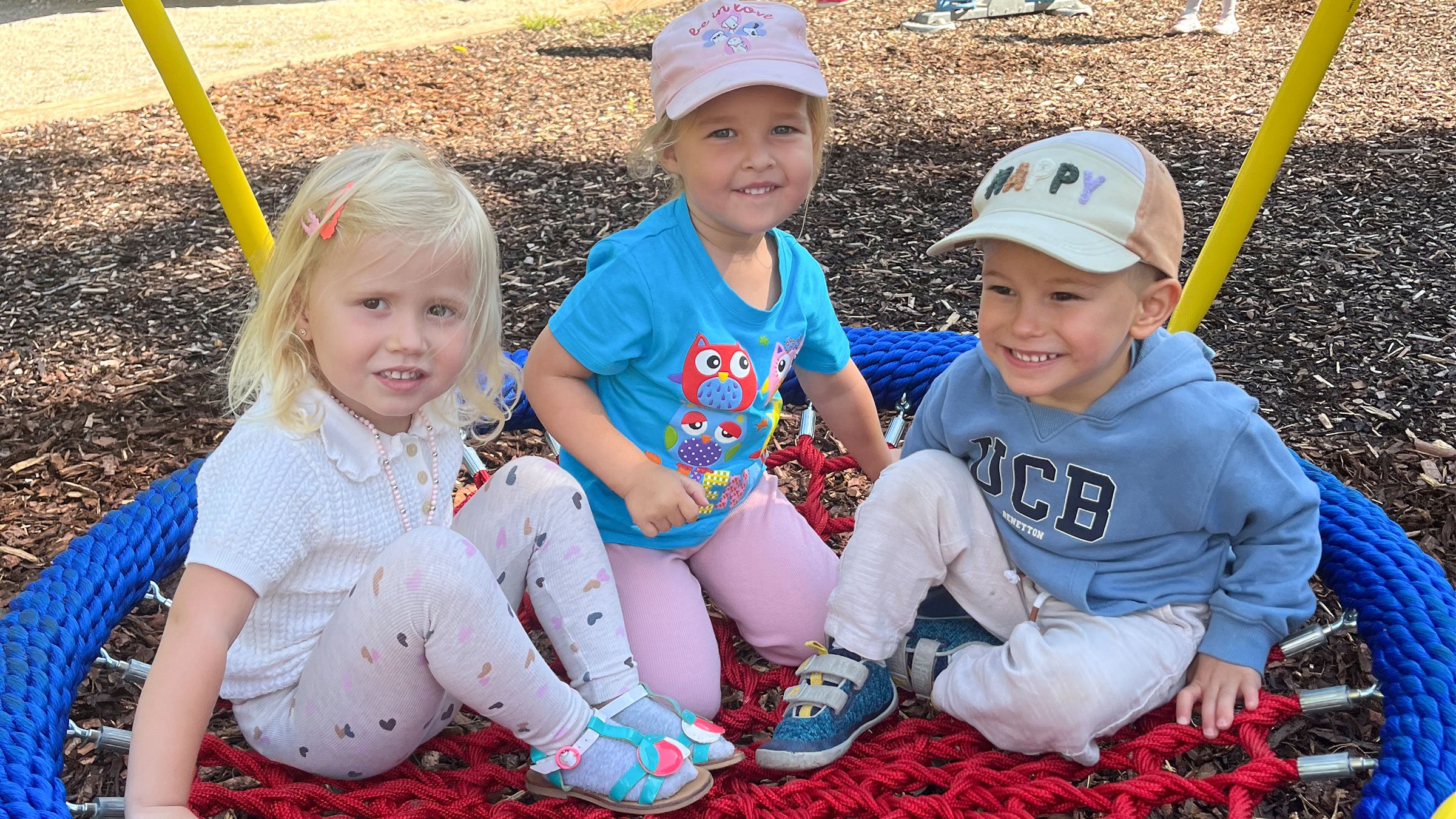 Drei kleinere Kinder mit Sommergewand bei Sonnenschein im Schatten in einer Nestschaukel sitzend.