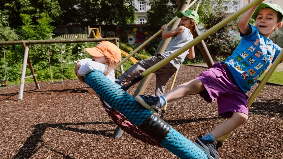 Drei Kinder in einer Netzschaukel auf einem weitläufigen Spielplatz mit Rindenmulch am Boden und Wiese und Bäumen im Hintergrund, zwei Buben stehend die Schaukel antreiben, ein Kind am Bauch in der Schaukel liegend.