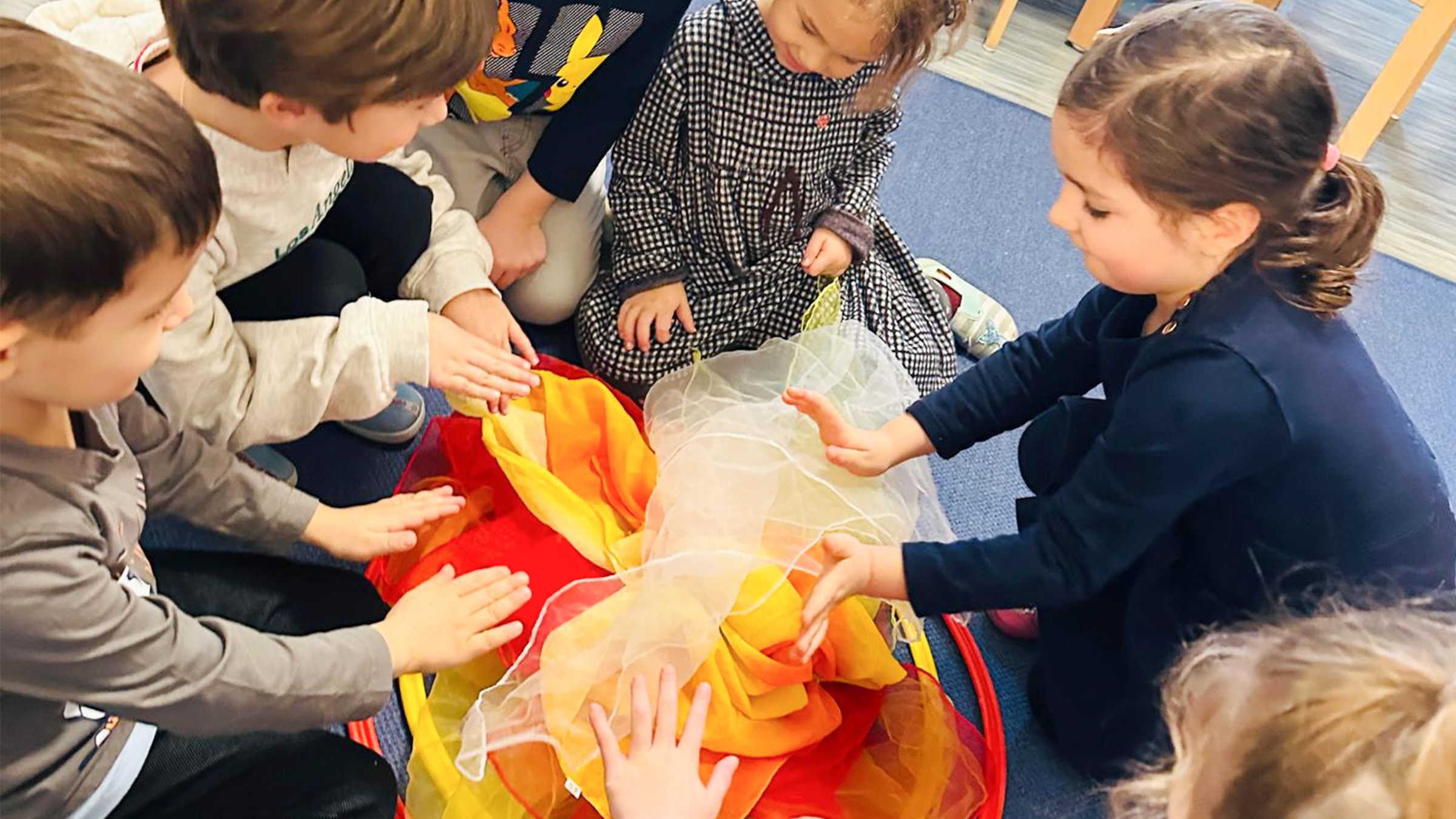 Gruppe von kleineren Kindern am Boden um rot, gelbe und orange Tücher sitzend.