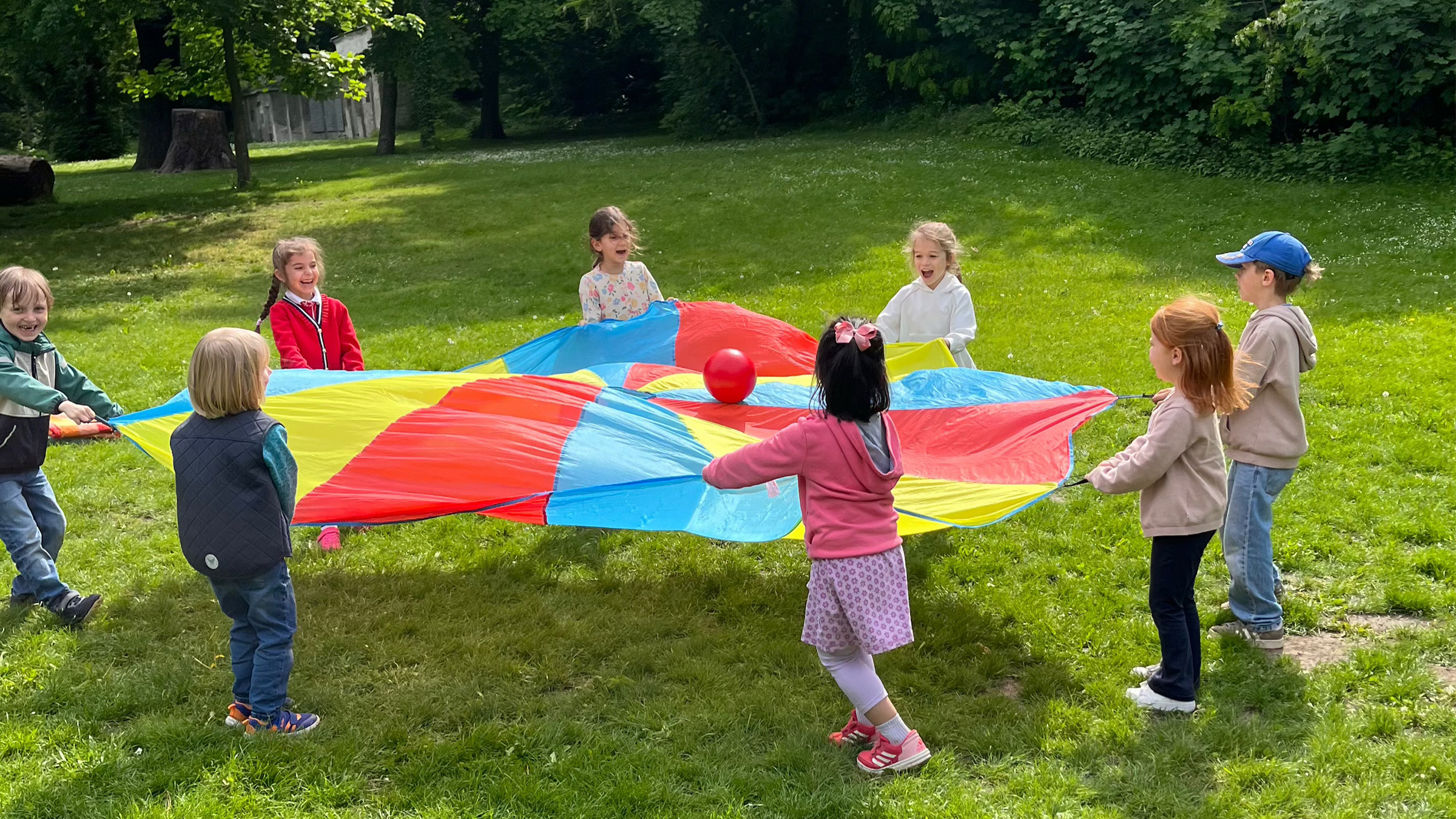 Gruppe von lachenden Kindern im Kreis auf einer Wiese mit Bäumen im Hintergrund, die einen roten Ball auf einem großem bunten Tuch zum Hüpfen bringen.