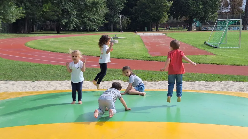 Fünf kleine Kinder auf einer Hüpfblase auf einem Sportplatz mit Leichtathletikbahn in sommerlicher Kleidung hüpfend und knieend.